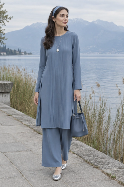 A woman wears a blue-gray long tunic with matching wide-leg trousers, a headband, pendant necklace, handbag, and silver flats beside a lakeside.