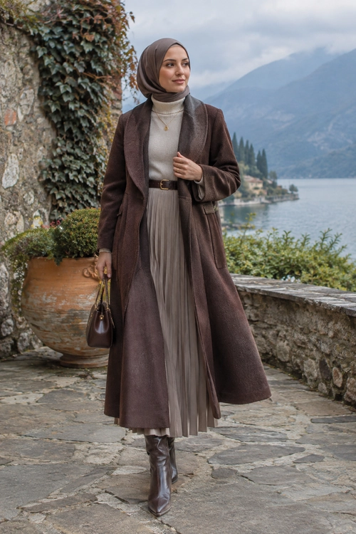 Elegant Brown Coat and Pleated Skirt A woman wears a long brown coat over a gray turtleneck and pleated skirt with a brown hijab, belt, handbag, and knee-high boots.