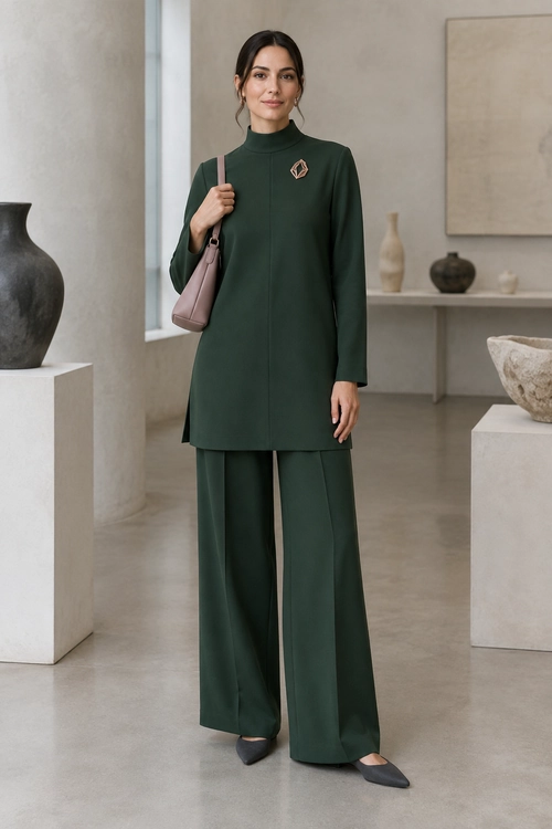 A woman wears a dark green high-neck tunic with wide-leg trousers, pointed flats, a mauve shoulder bag, and a brooch.