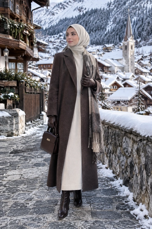 Elegant Winter Modest Outfit A woman wearing a long brown coat over a cream dress with a matching headscarf, fringed scarf, gloves, boots, and a structured handbag on a snowy village street.