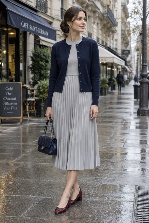 Navy Cardigan and Pleated Midi Dress Woman wearing a navy cardigan over a light gray pleated midi dress with burgundy pumps and a quilted chain-strap handbag.
