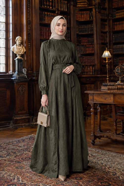 Elegant Olive Green Maxi Dress A woman wears a long olive green dress with a gathered waist, beige hijab, pointed shoes, and a small beige handbag.