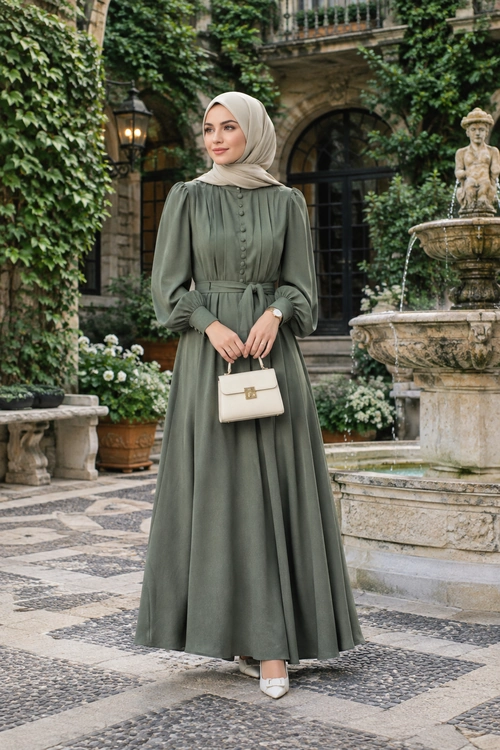 A woman wears an olive green layered maxi dress with a beige hijab, cream handbag, pendant necklace, and pointed shoes in a lush conservatory.