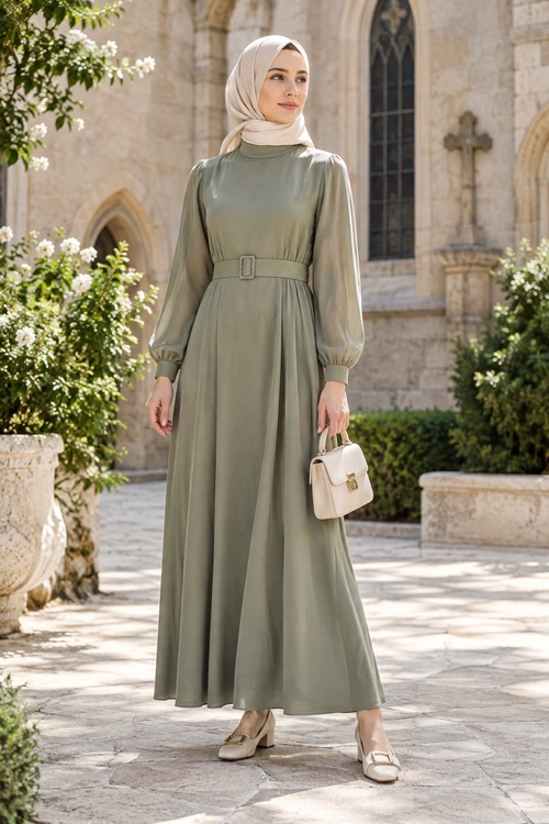 Woman wearing a sage green belted maxi dress with a cream hijab, structured handbag, and matching closed-toe shoes.