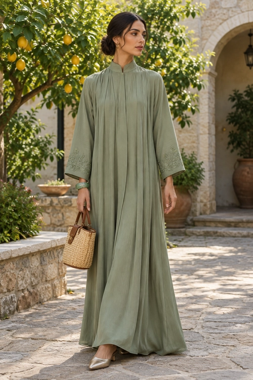 Sage Green Pleated Maxi Dress A flowing sage green pleated maxi dress with embroidered cuffs, a woven handbag, and pointed metallic shoes in a sunlit courtyard.