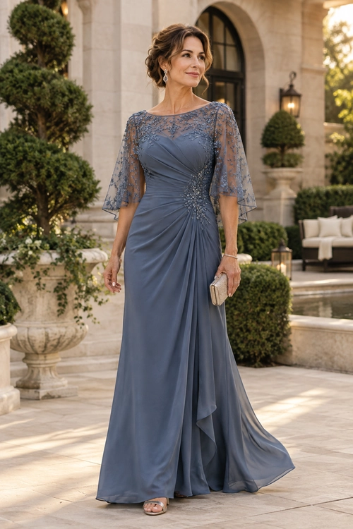 A woman wears a floor-length slate blue gown with embellished sheer cape sleeves, draped fabric, drop earrings, a small clutch, and open-toe sandals.