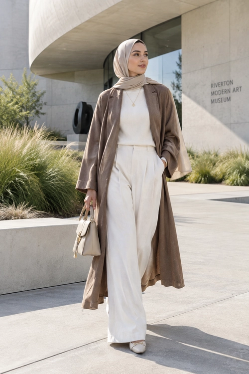 A woman wears a taupe hijab and long coat with a cream high-neck top, wide-leg trousers, handbag, and flats.